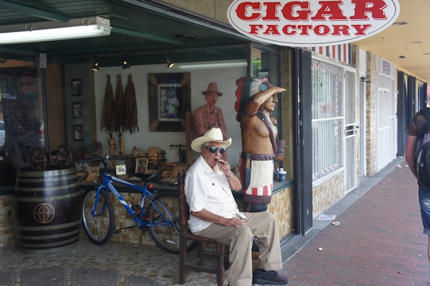 Cigar factory man sitting outside inviting folks in to pick up some cigars. Uncanny resemblance to the wood carving of a cigar smoking man on a chair in the glass display case.